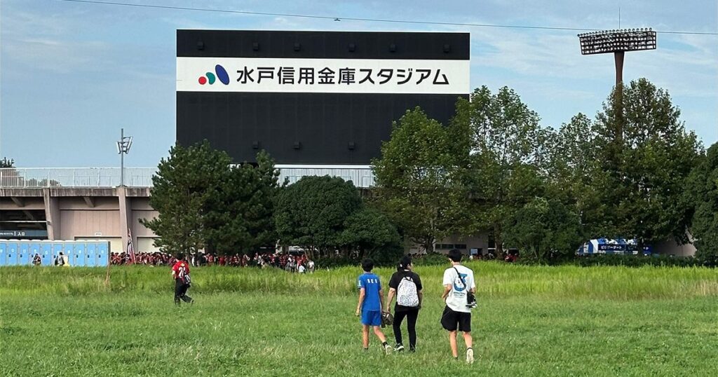 水戸信用金庫スタジアムの大型ビジョンと、手前の草地を歩いてスタジアムへ向かう来場者