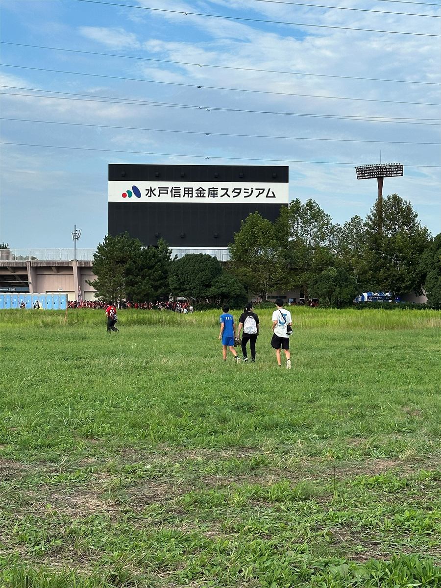 水戸信用金庫スタジアムへ草地を歩いて向かう来場者。奥に「水戸信用金庫スタジアム」と書かれた大型ビジョンが見える